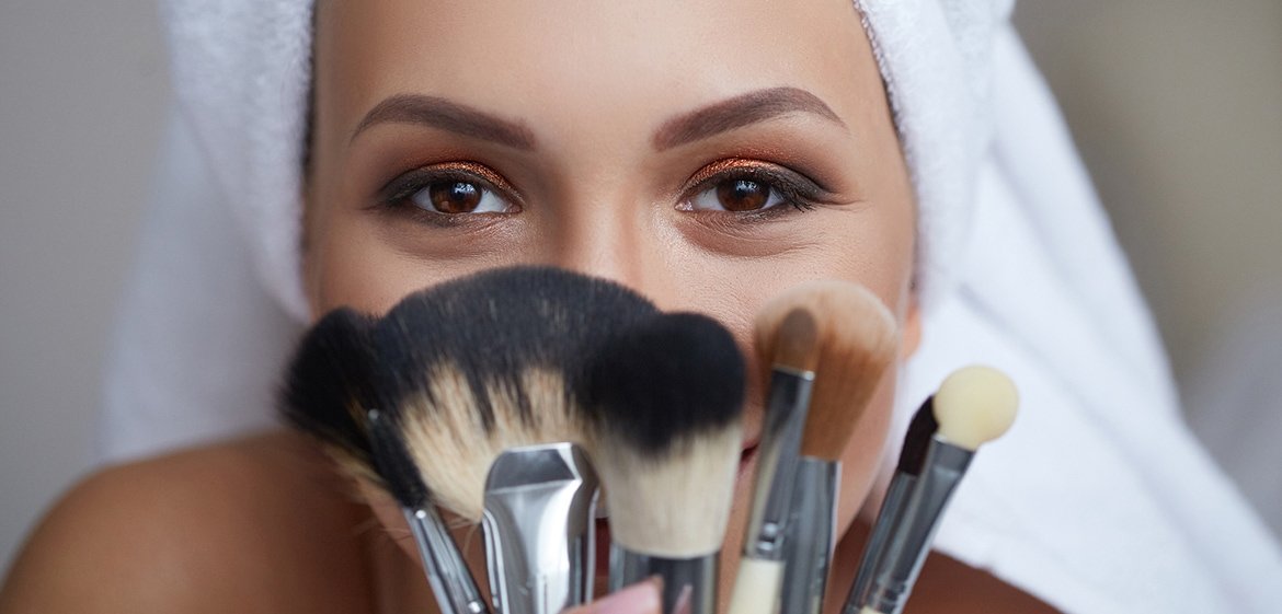 Concealer auftragen: Junge, geschminkte Frau hält eine Auswahl an Pinseln in die Kamera.